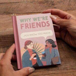 A small, pink and green fill-in-the-blank gift book titled 'Why We're Friends... You Know Too Much' being held open by a person's hands over a rustic wooden background. The cover features two illustrated women drinking coffee, emphasizing a close, humorous friendship. Ideal personalized gift for a best friend's birthday or Galentine's Day.