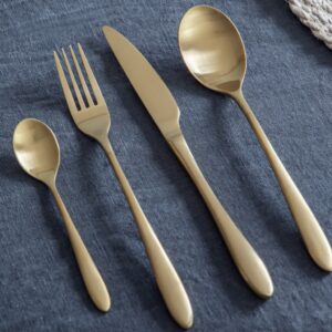 Four pieces of contemporary 16-piece cutlery set in a smooth, matte brass or brushed gold finish, including a fork, knife, dinner spoon, and teaspoon, resting on a dark blue linen tablecloth.