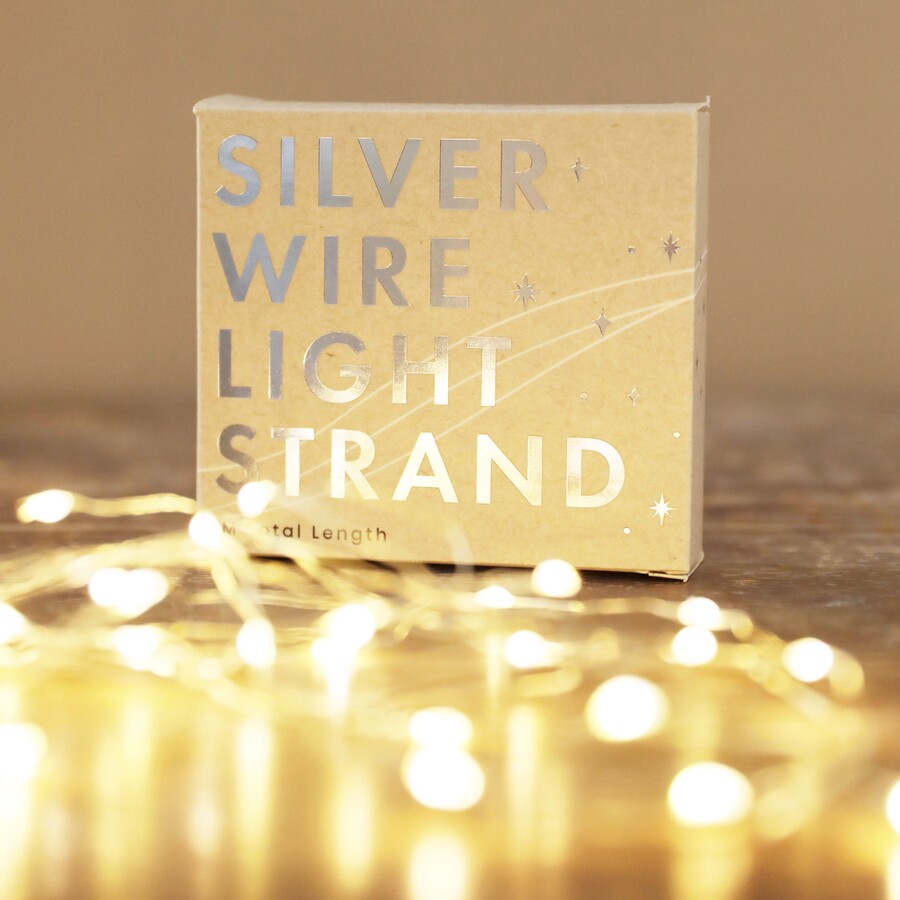 A small, gold-brown cardboard box for a light product, featuring the words "SILVER WIRE LIGHT STRAND" in reflective silver foil text, positioned on a wooden surface with a soft focus foreground of illuminated, warm-white LED lights on a thin silver wire.