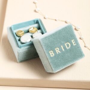 A small, square jewellery box with a hinged lid, upholstered in soft pale green velvet, featuring the word 'BRIDE' embossed in gold lettering on the lid, open to reveal a pair of white and gold stud earrings inside, with a pearl chain visible in the background.