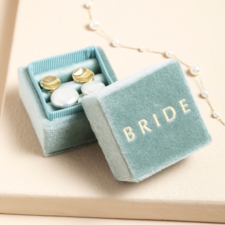 A small, square jewellery box with a hinged lid, upholstered in soft pale green velvet, featuring the word 'BRIDE' embossed in gold lettering on the lid, open to reveal a pair of white and gold stud earrings inside, with a pearl chain visible in the background.