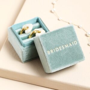 A small, square jewellery box with a hinged lid, upholstered in soft pale green velvet, featuring the word 'BRIDESMAID' embossed in gold lettering on the lid, open to reveal a pair of white and gold heart-shaped stud earrings inside, with a pearl chain visible in the background.