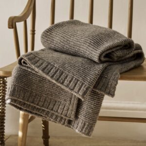Thick, chunky knit wool throw blanket in a natural taupe or brown-grey colour, featuring a seed stitch or moss stitch pattern and rib-knit border, folded over a wooden spindle chair.