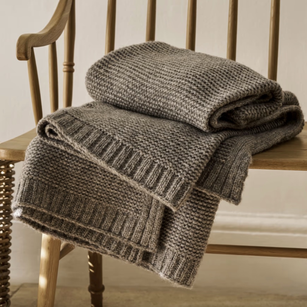 Thick, chunky knit wool throw blanket in a natural taupe or brown-grey colour, featuring a seed stitch or moss stitch pattern and rib-knit border, folded over a wooden spindle chair.