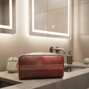 A luxury men's wash bag crafted from rich, reddish-brown Buffalo leather, featuring three horizontal rows of white stitching designed to resemble a cricket ball seam, sitting on a modern grey stone bathroom counter.