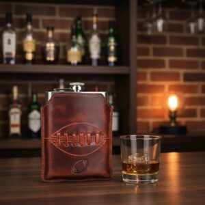 Stainless steel hip flask encased in brown buffalo leather with detailed rugby ball stitching, sitting on a wooden bar counter.