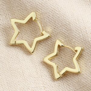 Close-up of a pair of gold-plated open star-shaped hoop earrings with a polished finish, resting on a neutral, textured beige fabric, highlighting the modern, celestial design.