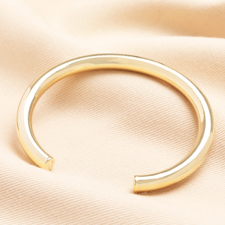 A simple, thick gold-toned open torque bangle with a smooth, highly polished finish, resting on a textured pale beige fabric surface with its open ends facing slightly upwards.