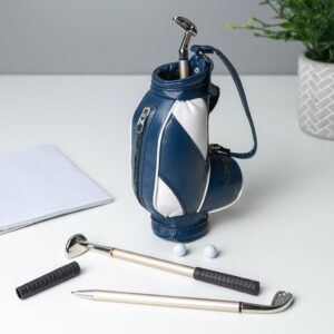 A blue and white miniature golf bag standing on a white desk next to a notebook. In front of the bag are two golf pens, one without a cap showing the metal tip, and two tiny white golf balls.