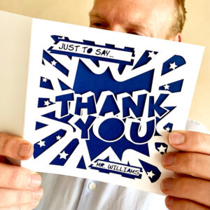 A man holding a personalised laser-cut thank you card with a comic book 'POW!' design, featuring a navy blue insert with 'THANK YOU' and a custom name ('Mr. Williams') in bold lettering.
