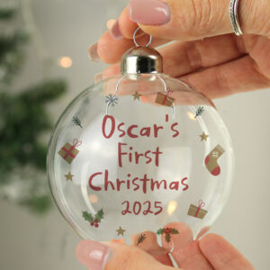 A hand holding a clear glass Christmas bauble with a chrome cap, personalized with the name Oscar's and the year 2025 in red vinyl, surrounded by festive icons like stars, gifts, and holly.