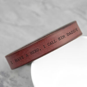 Close-up of a thick, personalized brown leather cuff bracelet with a custom engraved message, 'I HAVE A HERO, I CALL HIM DADDY', resting on a white surface.