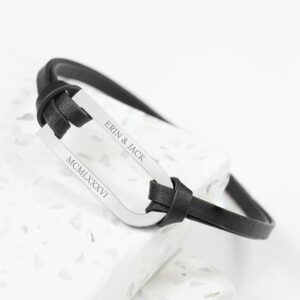 Close-up photograph of a personalised men's silver bar leather bracelet. The bracelet features a polished silver bar, engraved on the visible side with "ERIN & JACK" on the top line and the Roman numerals "MCMLXXXVI" on the bottom line. The silver bar is threaded through a thin black leather strap, which is tied in knots at both ends of the bar. The bracelet is displayed on a white, speckled surface.
