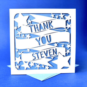 Close-up of a personalised thank you card featuring white laser-cut paper with large banners reading 'Thank You' and a custom name (Steven), surrounded by scattered stars, backed by a light blue insert.