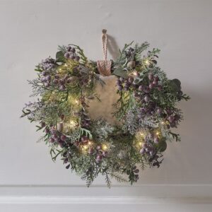 Decorative heart-shaped Christmas wreath featuring frosted purple berries, mixed artificial pine and cedar-like greenery, and warm white micro fairy lights, tied with jute rope and hanging on a pale wall.