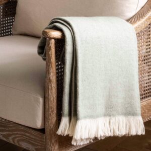 Luxury wool throw blanket in a pale sage green herringbone pattern with a thick cream fringe, folded and draped over the arm of a wooden armchair with cane webbing, creating a cosy Scandi look.