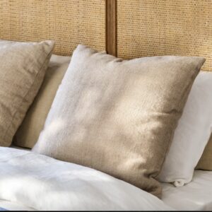 Square accent cushion in a natural oatmeal or light beige color with a textured herringbone woven pattern, resting on white bedding against a rattan cane headboard.