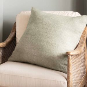 Square accent cushion in a muted sage green color with a visible subtle herringbone woven pattern, resting on a beige armchair with a natural cane frame.
