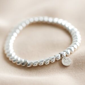 A delicate silver stretch bracelet composed entirely of interlocking, smooth silver heart-shaped beads, featuring a small, round silver charm with a subtle inscription hanging near the clasp, displayed on a textured pale fabric background.