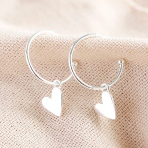 A pair of simple, polished silver hoop earrings, each featuring a dangling, solid silver heart charm hanging slightly askew, displayed against a textured pale beige fabric background.
