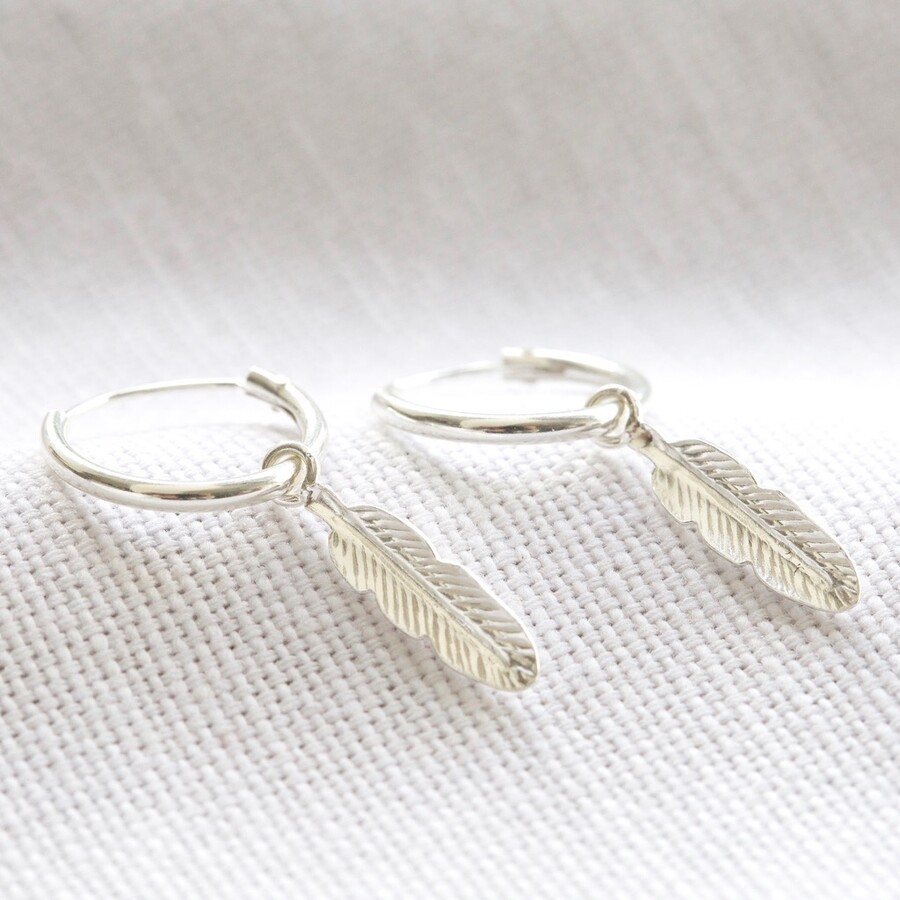 A pair of small, polished sterling silver hoop earrings, each adorned with a delicate, intricately detailed sterling silver feather charm dangling from the base, displayed against a soft, neutral-colored fabric background.