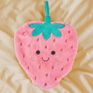 A smiling pink fluffy strawberry hot water bottle cover with green leaves and a handle, resting on a soft yellow blanket or duvet in a cosy bedroom scene.