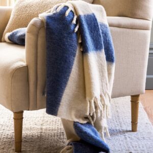 Thick, luxury 100% New Zealand Wool throw blanket in a wide stripe pattern of deep indigo blue and natural cream, finished with a fringe, draped over a neutral armchair.