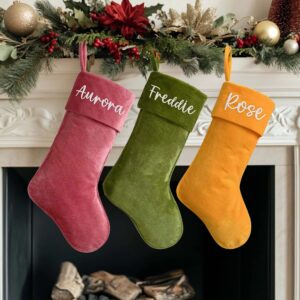 Three luxury personalised velvet Christmas stockings in deep festive colours—pink, green, and orange/yellow—each embroidered with a white script name ('Aurora', 'Freddie', 'Rose'), hanging from a white fireplace mantelpiece decorated with artificial pine garlands and red berries.