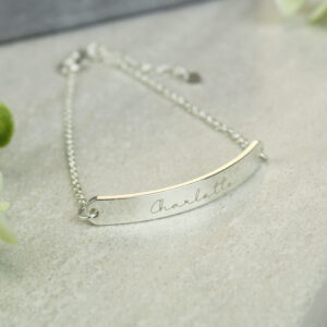 Close-up of a delicate silver chain bracelet with a curved central bar, engraved with the cursive name 'Charlotte', resting on a light grey stone surface with soft light reflecting on the polished metal.