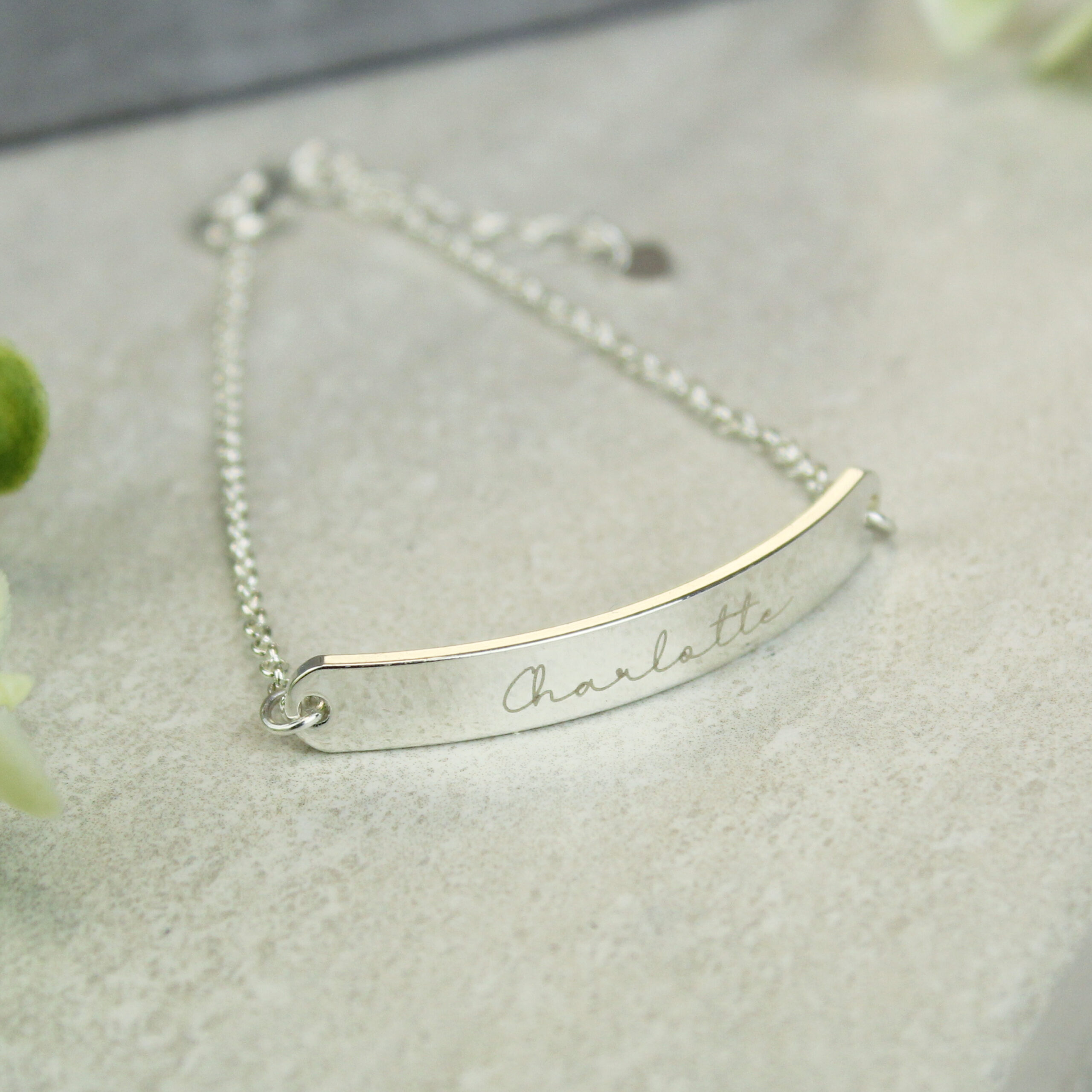 Close-up of a delicate silver chain bracelet with a curved central bar, engraved with the cursive name 'Charlotte', resting on a light grey stone surface with soft light reflecting on the polished metal.