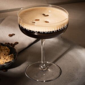 A wide, stemmed coupe glass filled with a dark espresso martini cocktail, topped with a rich layer of foam and three coffee beans, featuring the personalised engraved text 'ESPRESSO MARTINI CLUB' wrapped around the rim.
