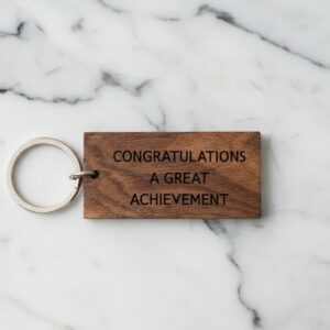 Personalised rectangle keyring made from dark walnut wood, engraved with the text 'CONGRATULATIONS A GREAT ACHIEVEMENT', attached to a metal split ring, displayed on a white marble surface.