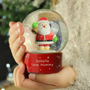 A child's hands holding a Personalised Santa Snow Globe with a bright red base, featuring a classic Santa figure holding a bell inside, and the custom engraved message 'Isabelle Love Mummy' on the base with white star details.