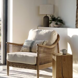 Natural wooden armchair with rattan cane detailing, thick off-white linen cushions, and a small decorative striped pillow, placed in a bright, modern living room.