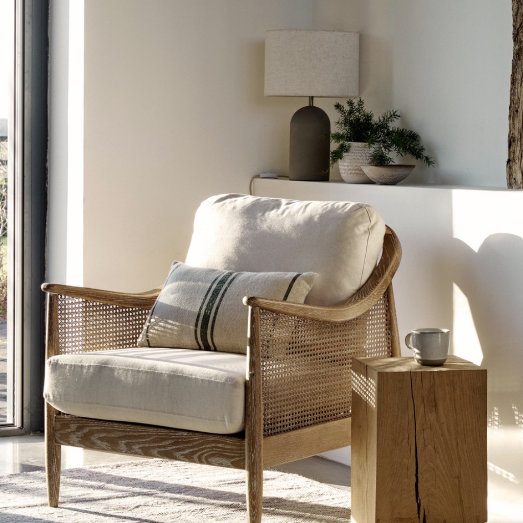 Natural wooden armchair with rattan cane detailing, thick off-white linen cushions, and a small decorative striped pillow, placed in a bright, modern living room.