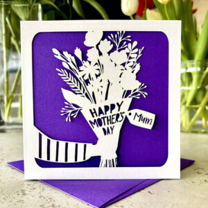 A luxury laser-cut Mothering Sunday card featuring a white paper-cut silhouette of a hand holding a flower bouquet against a vibrant purple background, with a personalised name tag that says "Mum".