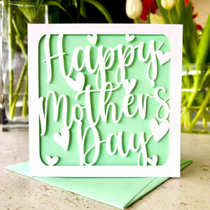 A luxury laser-cut Mother's Day card featuring "Happy Mother’s Day" in white script with delicate heart cut-outs, set against a soft mint green background with a matching mint envelope.