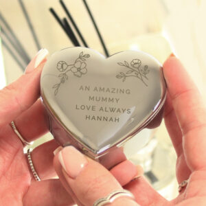 A pair of hands holding a polished silver heart-shaped trinket box. The lid is engraved with delicate floral illustrations in the top corners and a personalised message in the centre that reads: "AN AMAZING MUMMY LOVE ALWAYS HANNAH." The background is softly blurred, showing a warm, indoor setting with a reed diffuser.
