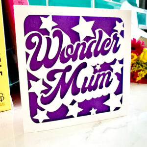 A luxury square Mother’s Day card featuring the words "Wonder Mum" in a bold, white cursive laser-cut font. The design is surrounded by white star cut-outs, all layered over a vibrant, textured purple background.