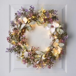 A large 60cm spring wreath with cream, lilac, and rose flowers and sage green leaves, lit with warm white fairy lights on a white door.