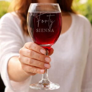 A hand holding a large, elegant wine glass filled with red wine. The glass is custom engraved with "Forty" in a delicate script font and "SIENNA" in bold uppercase letters. A soft-focus outdoor background highlights the crystal-clear glass.