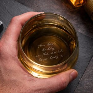A hand holding a weighted tumbler filled with an amber-coloured spirit, looking down through the glass to reveal the base-engraved message 'the best daddy in the world love lucy x'.