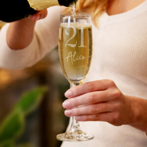 A person in a white top holding a champagne flute while sparkling wine is poured from a bottle. The glass is engraved with a large number '21' and the name 'Alice' in a stylish script font