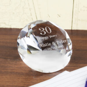 A brilliant-cut clear glass diamond paperweight resting on a wooden surface. The flat face is engraved with a large "30" at the top, followed by "Happy Years" and the names "Katie & Peter" in a larger script font, finished with the date "05/05/2015" at the bottom.