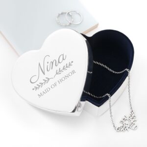 Top-down view of a heart-shaped silver jewellery box with a navy velvet lining. The lid is engraved with "Nina" in a script font and "Maid of Honor" in a serif font, separated by a delicate floral leaf design. A silver bee necklace is draped inside.
