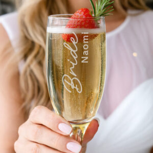 A close-up of a person holding an elegant champagne flute filled with sparkling wine, garnished with a strawberry and rosemary. The glass is engraved with 'Bride' in large script and the name 'Naomi' in vertical block text.