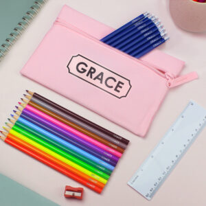 A pink microfibre pencil case with the name "GRACE" printed in a bold black frame. Next to the case is a set of multi-coloured crayons and blue pencils, all individually personalised with the name "GRACE". The set is arranged on a desk with a white ruler and a red pencil sharpener.