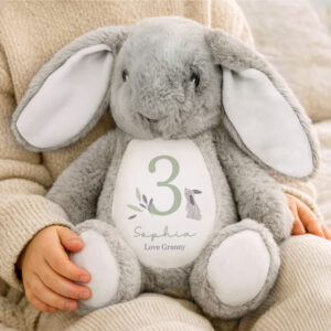 A child sitting in a cream knitted jumper holding a soft grey plush bunny. The bunny has long floppy ears and a white belly featuring a sage green number '3', the name 'Sophia' in script, and 'Love Granny' printed below. The design is accented with small lilac botanical leaves and a tiny illustrated rabbit.