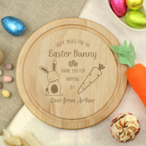 A round wooden treat board for the Easter Bunny, featuring engraved illustrations of a rabbit and a carrot. The board is personalised at the bottom with the text "Love from Arthur". It is styled on a white marble surface surrounded by colourful foil-wrapped chocolate eggs and a felt carrot toy.
