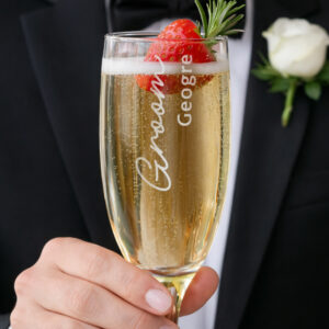 A close-up of a person in a black tuxedo and bow tie holding a champagne flute filled with bubbly, garnished with a strawberry and rosemary. The glass is engraved with 'Groom' in script and the name 'George' vertically.